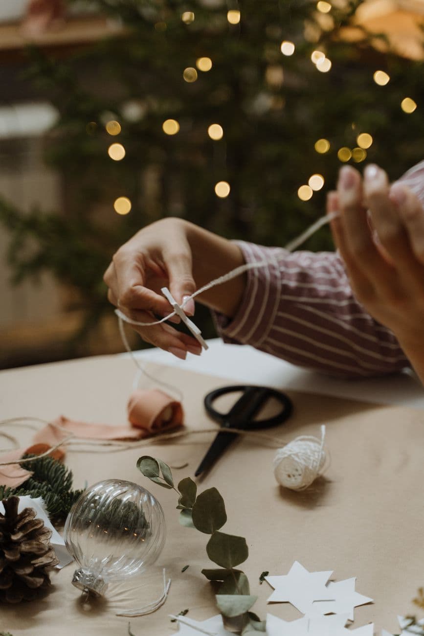 Hands making Christmas decorations using pine cones and stars inside a cozy home.