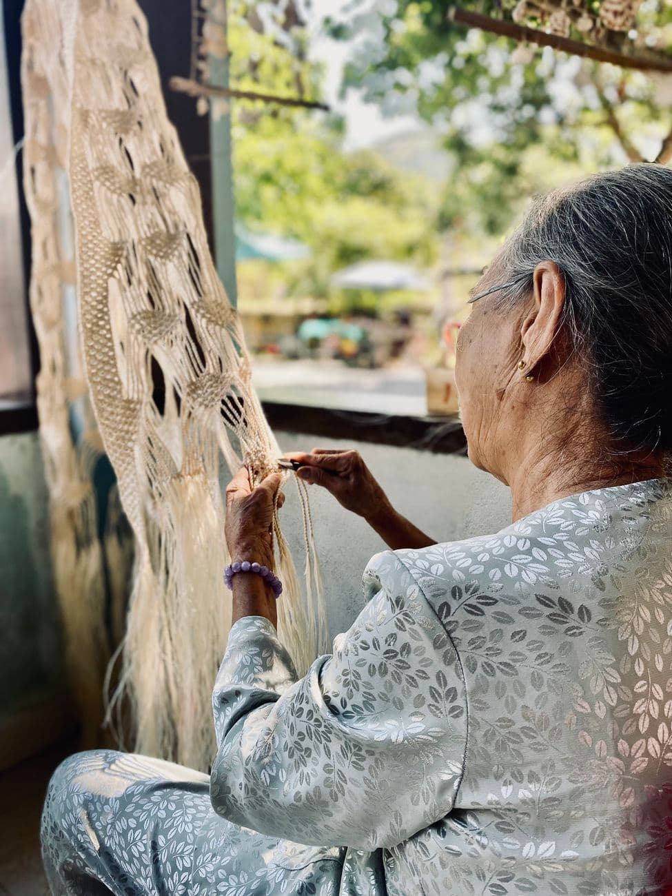Senior woman skillfully creating macrame art outdoors on a sunny day, demonstrating traditional craftsmanship.